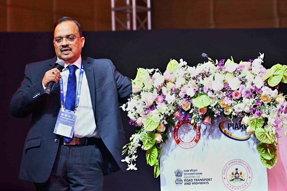 Dr Hegde speaking into a microphone on stage beside a flower-decked lectern bearing the Indian Roads Congress emblem